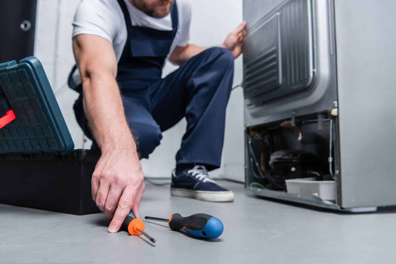 Technician kneeling on the floor with tools while repairing the back of a refrigerator.